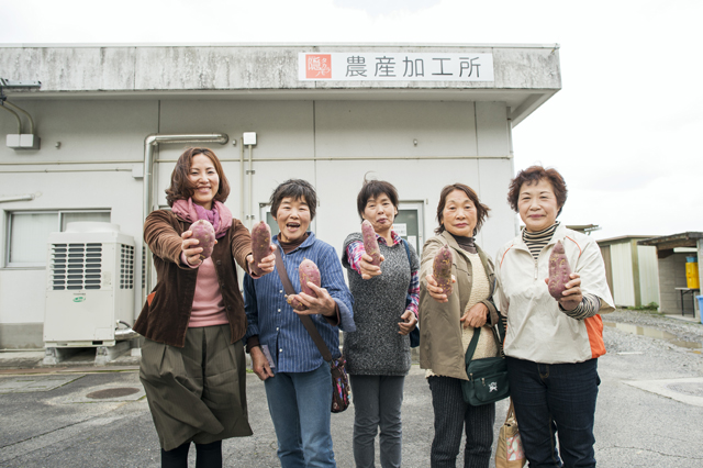 Ms. Sugioka and some farmers holding sweet potatoes and smiling in front of the factory